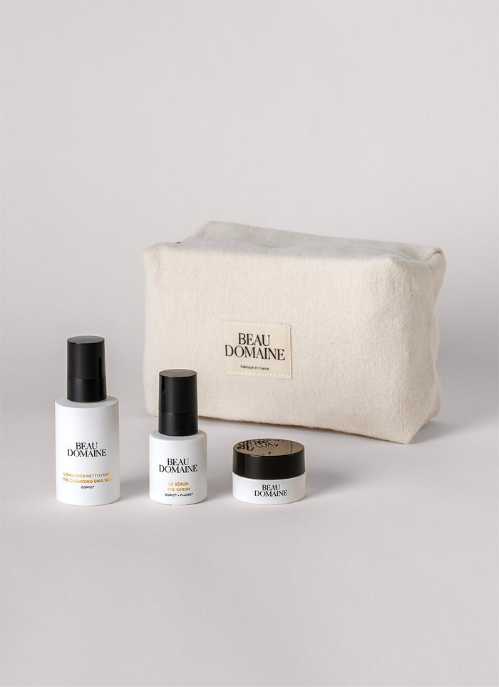Conjunto cosmético com frascos e um frasco de creme à frente de uma pochette 'Beau Domaine' sobre fundo cinzento claro